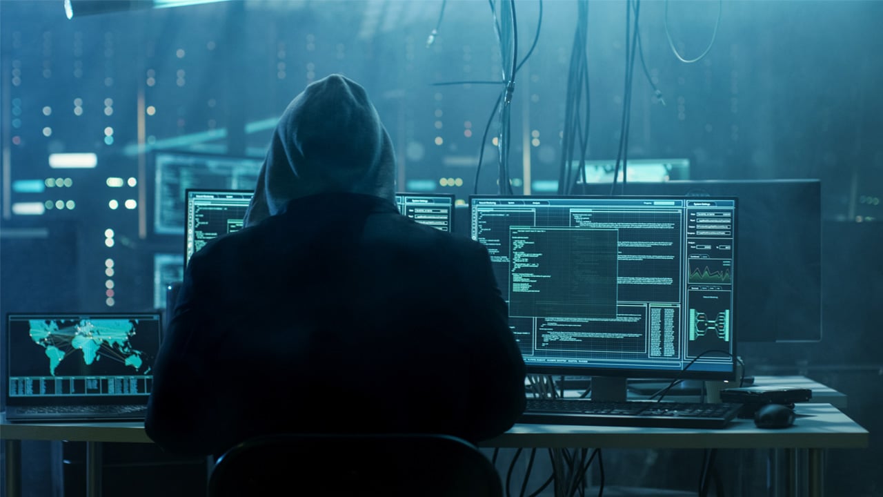 A person wearing a hoodie sits at a desk, focused on multiple computer monitors.