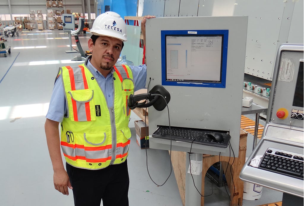 A Tecesa Architectural Glass employee wearing a white hard hat and a high-visibility vest stands next to a workstation featuring a computer monitor displaying FeneVision software. The background shows an industrial glass manufacturing facility.