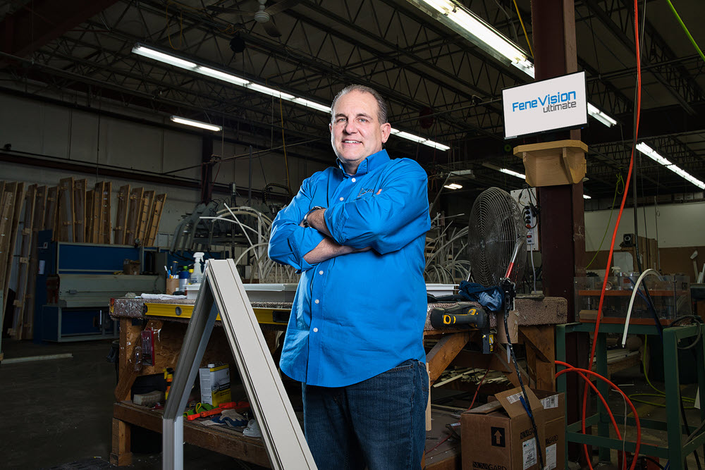 A Fen-Tech employee in a blue company shirt standing confidently with arms crossed in a factory setting. Various tools, equipment, and a FeneVision Ultimate sign are visible in the background.