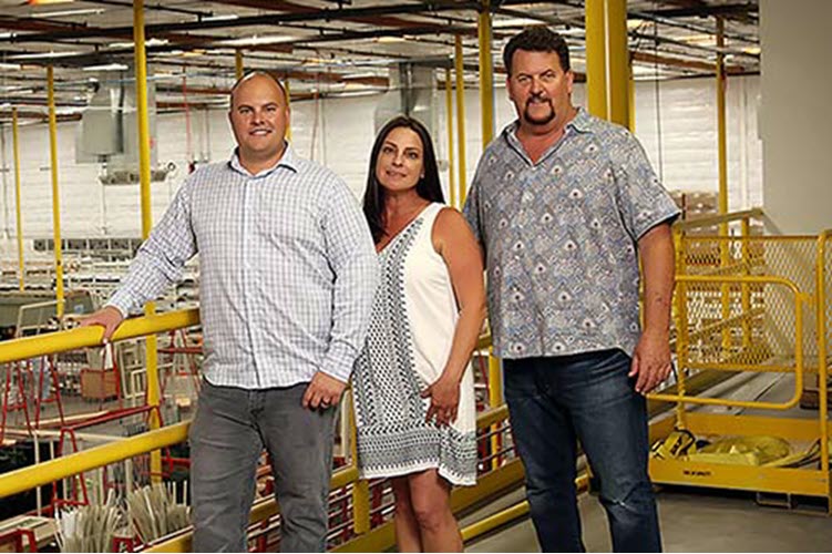 The Avanti management team standing in a warehouse with yellow railings: a bald man in a checkered shirt, a woman in a white dress, and a man in a blue patterned shirt.