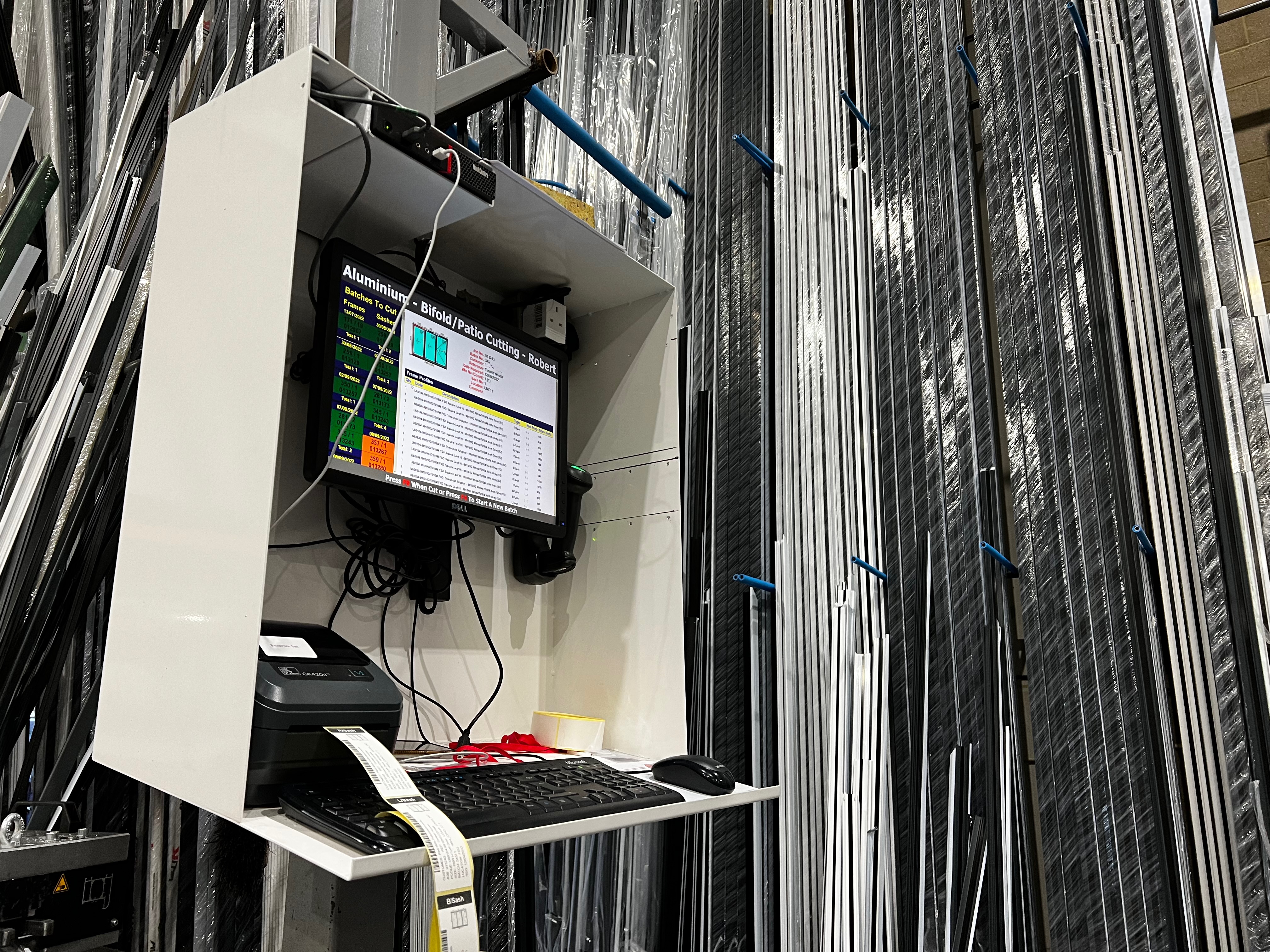 A workstation in a workshop with a computer screen displaying job information for aluminum bifold/patio cutting. A label printer and keyboard are on the desk, with metal profiles stored vertically in the background.