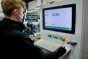 A worker wearing earplugs operates a control panel with a large display screen in a manufacturing setting. The screen shows a schematic and process information. The background includes various industrial equipment and materials.