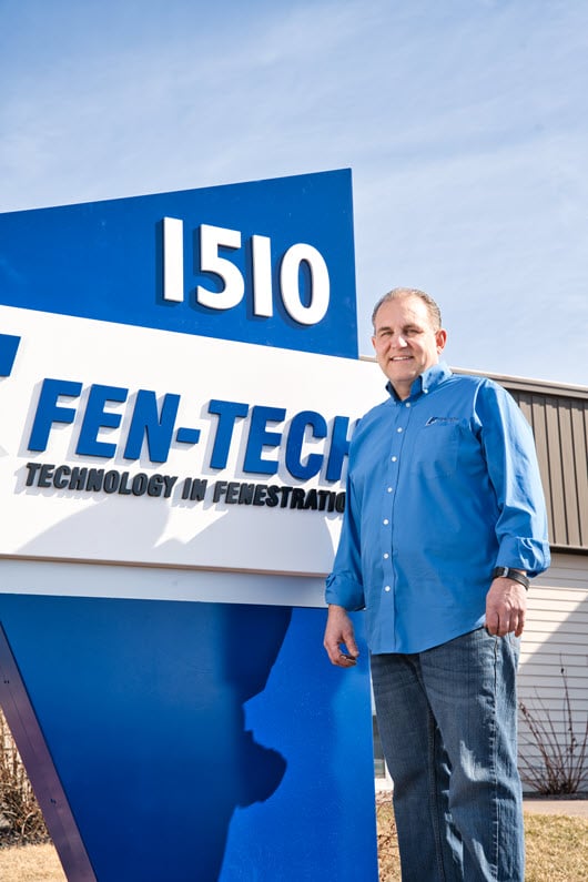 A Fen-Tech employee standing outside next to a large Fen-Tech sign that reads 'Technology in Fenestration' and the address number 1510. The employee is wearing a blue Fen-Tech shirt and smiling at the camera."