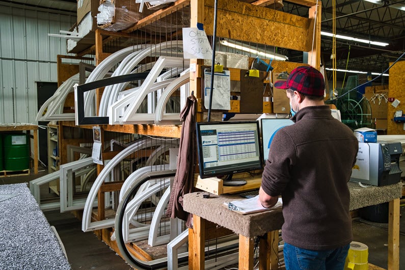 A Fen-Tech employee wearing a red and black checkered cap, standing at a computer station in a warehouse. The workstation has a monitor displaying data, and shelves filled with window frames are visible in the background."