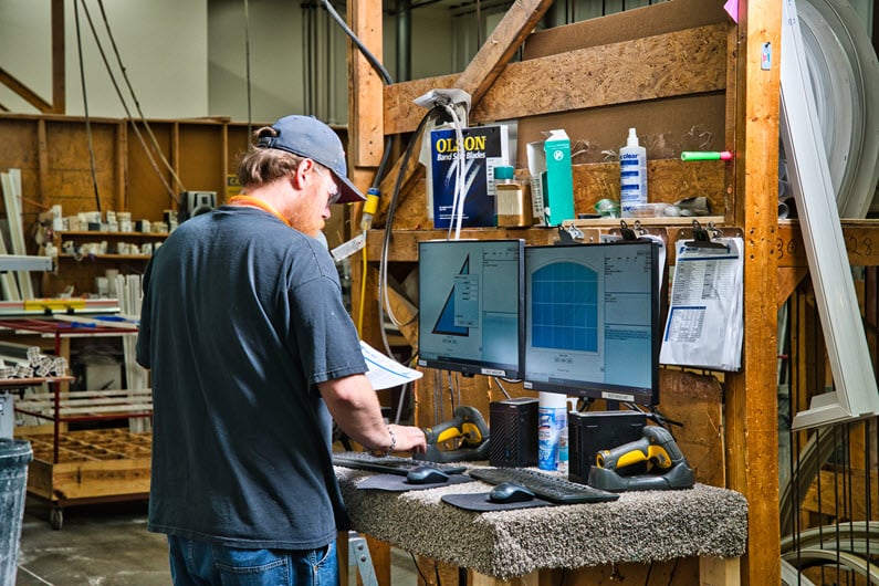 A Fen-Tech employee wearing a cap and safety glasses working at a computer station in a workshop. The workstation has dual monitors displaying design specifications and various tools and supplies are visible around the desk.