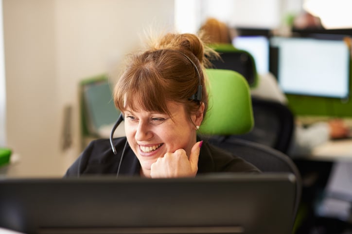 technical support representative with reddish hair tied in a bun is smiling while wearing a headset, engaged in a conversation. She is sitting in a modern office environment with green ergonomic chairs and other employees working at their desks in the background.