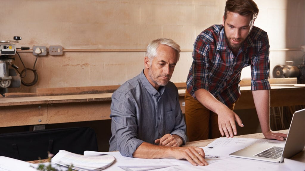 Two men collaborating on a laptop in a woodworking shop.