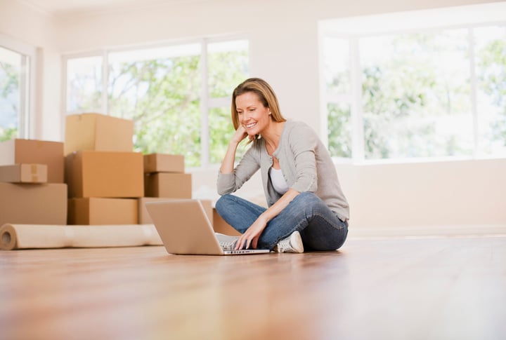 A woman sitting on the floor with a laptop, smiling as she works. The room is brightly lit with large windows, and there are moving boxes and a rolled-up carpet around her.