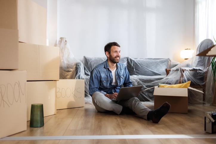 A man sitting on the floor surrounded by moving boxes, working on a laptop. The room is sparsely furnished with a couch covered in protective plastic.