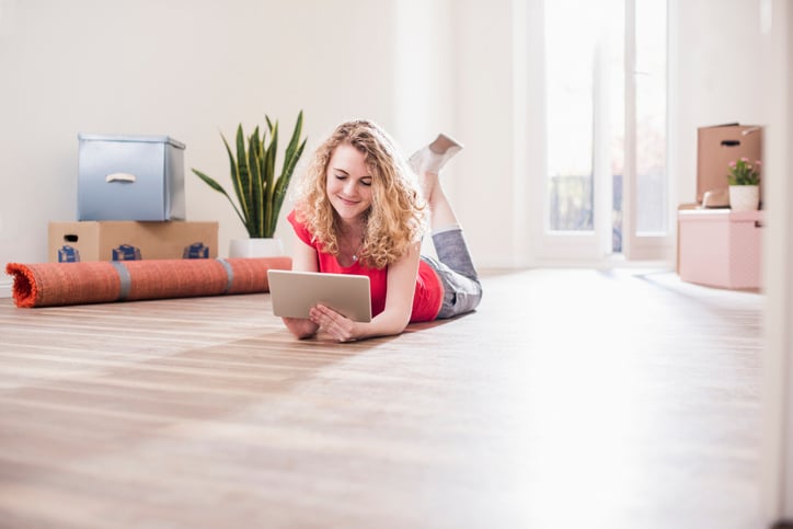 A woman lying on the floor, using a tablet. The room is filled with moving boxes, a rolled-up carpet, and plants, and is lit by natural light from a nearby window.