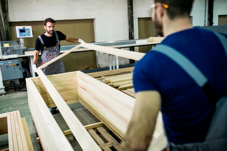 Two workers in a woodworking shop assembling a large wooden frame, wearing safety gear and handling the materials carefully.