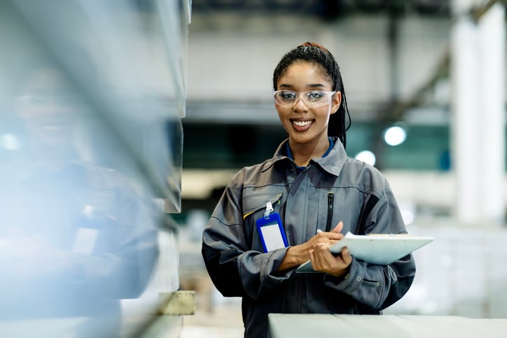 A smiling female factory worker wearing safety goggles and a gray uniform, holding a clipboard in an industrial setting.