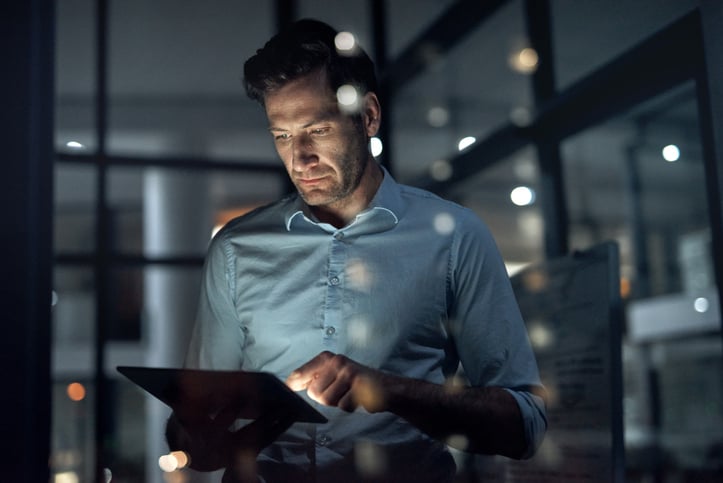 A man in a dimly lit office working on a tablet, with city lights reflecting in the glass behind him.