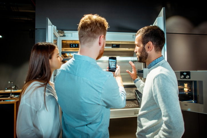 Three people reviewing appliance details on a smartphone while standing in front of an open kitchen appliance display.