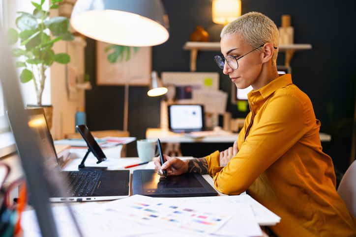 A designer working on a digital tablet at her desk, surrounded by design tools and a laptop, in a well-lit, modern home office.