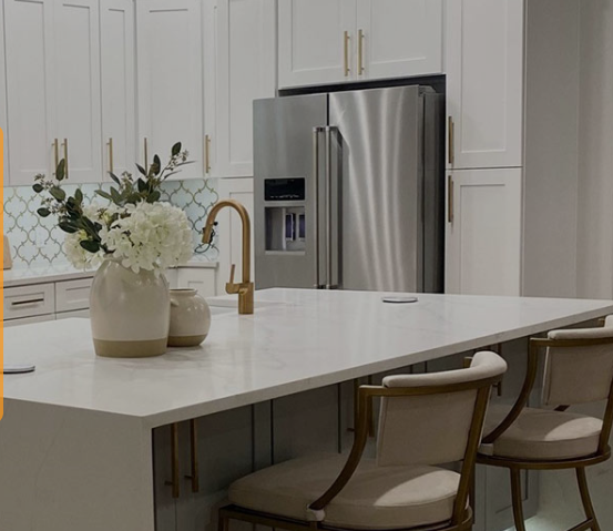 A modern kitchen with white cabinets and gold hardware, a large marble island with decorative vases and seating, complemented by a stainless steel refrigerator and geometric backsplash tiles.