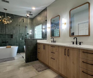 A modern bathroom design featuring a double vanity with light wood cabinetry, black fixtures, and rectangular mirrors framed in wood. The space includes a luxurious walk-in shower with floor-to-ceiling dark green tiles and a freestanding bathtub under a gold chandelier, creating a sleek and elegant atmosphere.