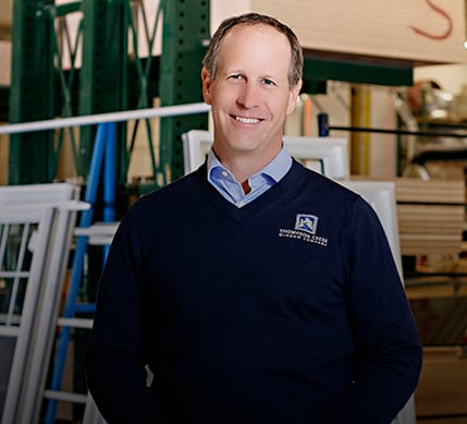 A Thompson Creek representative in a navy blue sweater with the company logo smiles while standing in a window production area, with stacks of frames and a worker assembling windows in the background.