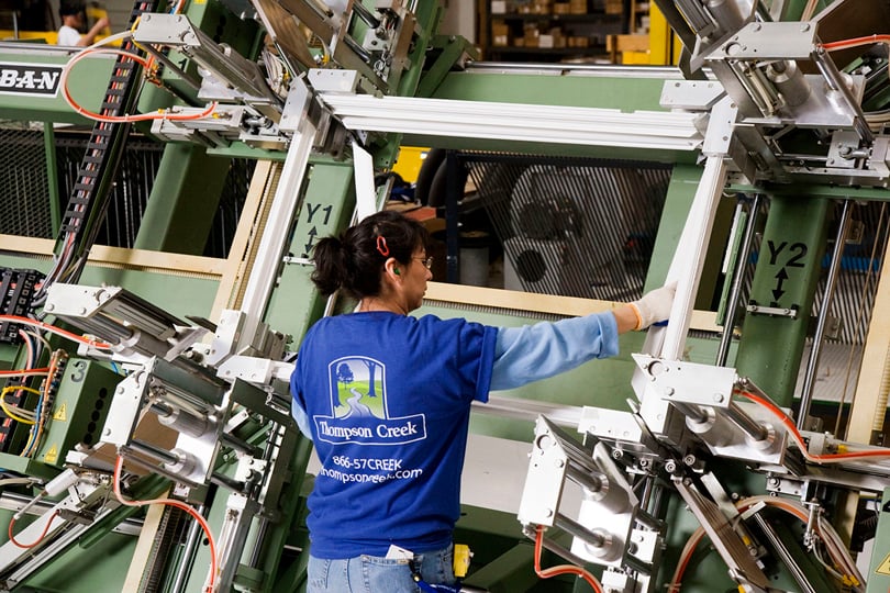 A Thompson Creek employee wearing a branded blue T-shirt assembles a window frame using advanced manufacturing equipment. The industrial setting features machinery and tools for precise assembly.