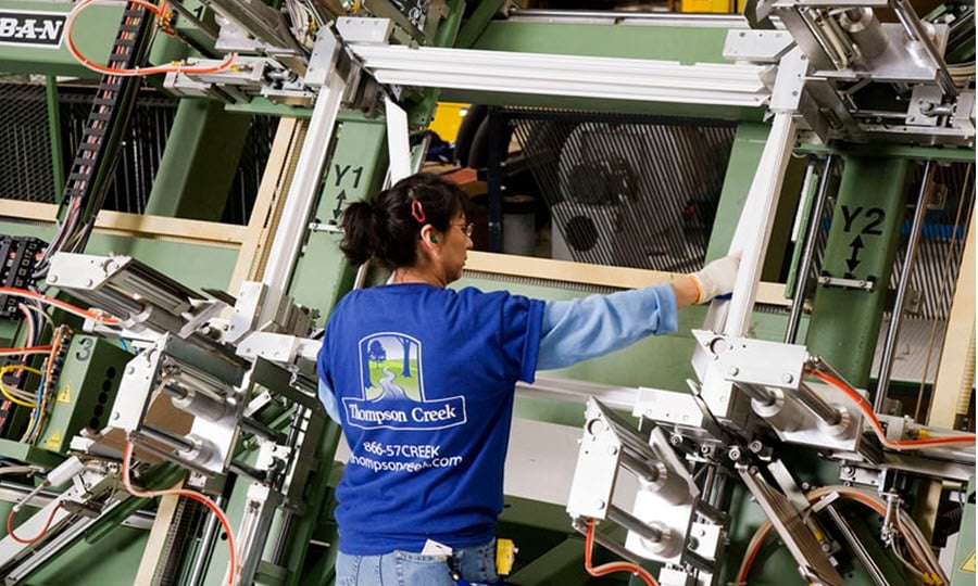 A Thompson Creek employee wearing a branded blue T-shirt assembles a window frame using advanced manufacturing equipment. The industrial setting features machinery and tools for precise assembly.