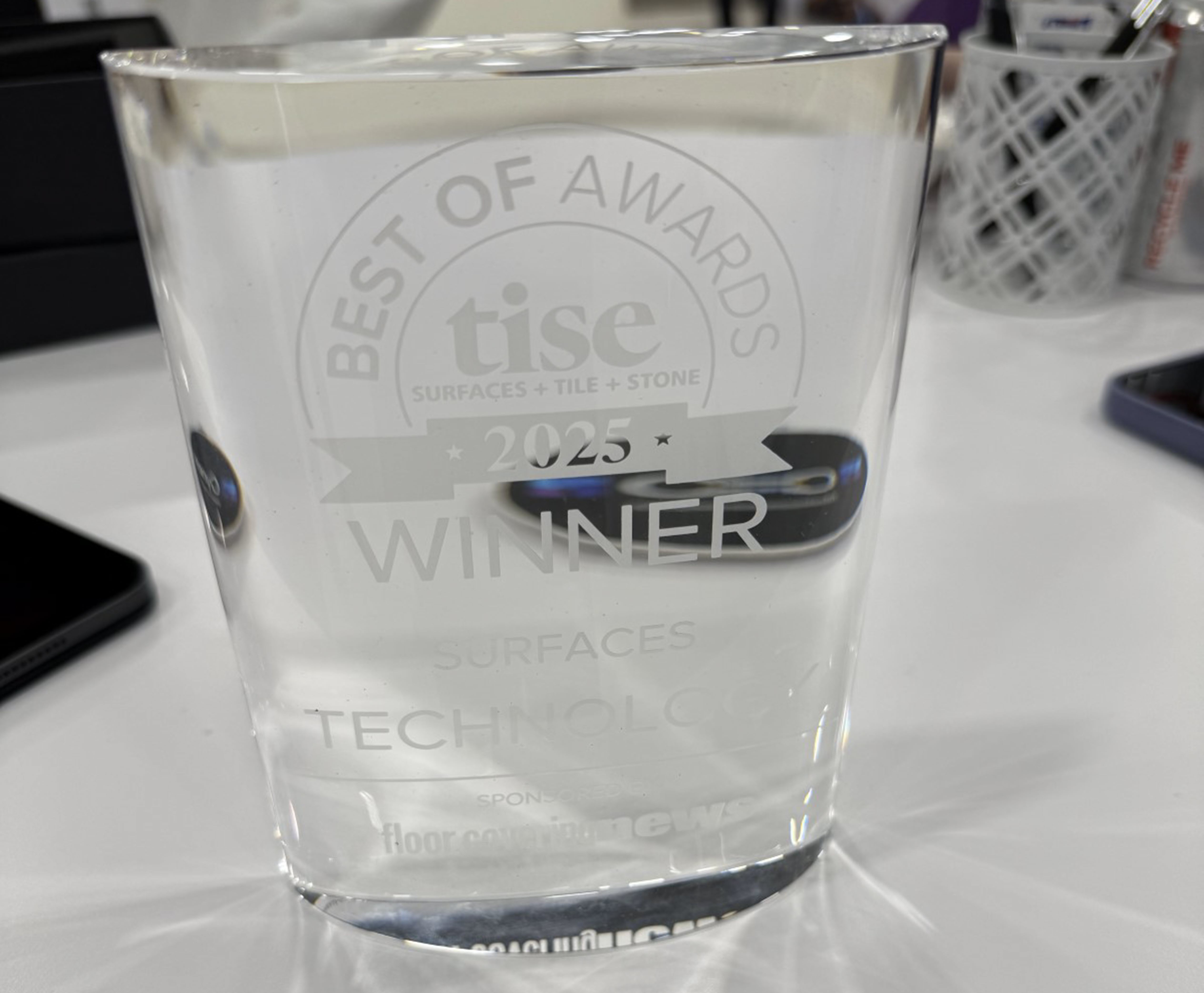 A clear, rectangular glass award with engraved text reading 'Best of Awards - TISE Surfaces + Tile + Stone - 2025 Winner - Surfaces Technology,' placed on a white desk. The award is sponsored by Floor Covering News. A reflection of a smartphone is visible on the surface of the award.