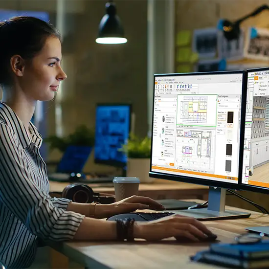 A woman sits at a desk in a modern office, working on a kitchen design project using dual monitors displaying CAD software. The environment is dimly lit with desk lamps, and she appears focused on her work.