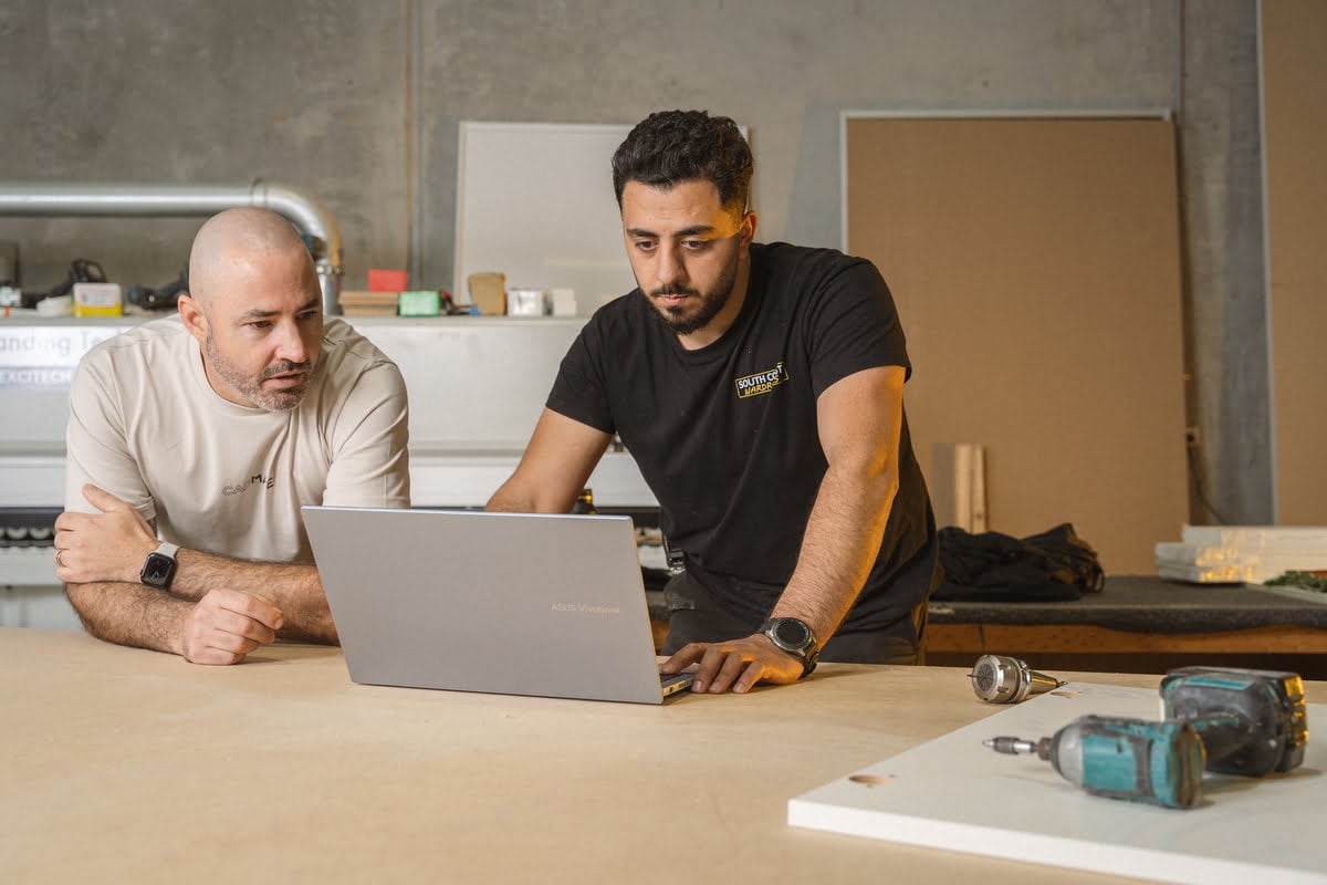 Two men collaborate on a laptop in a woodworking shop